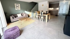 Un salon moderne avec un canapé, une table basse et un coin repas. On aperçoit une cuisine à l’arrière-plan, avec des placards et un réfrigérateur.