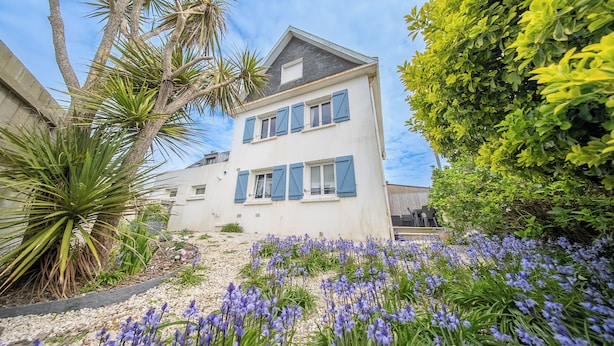 A two-story building with a white facade and blue shutters, surrounded by lush greenery and purple flowers.