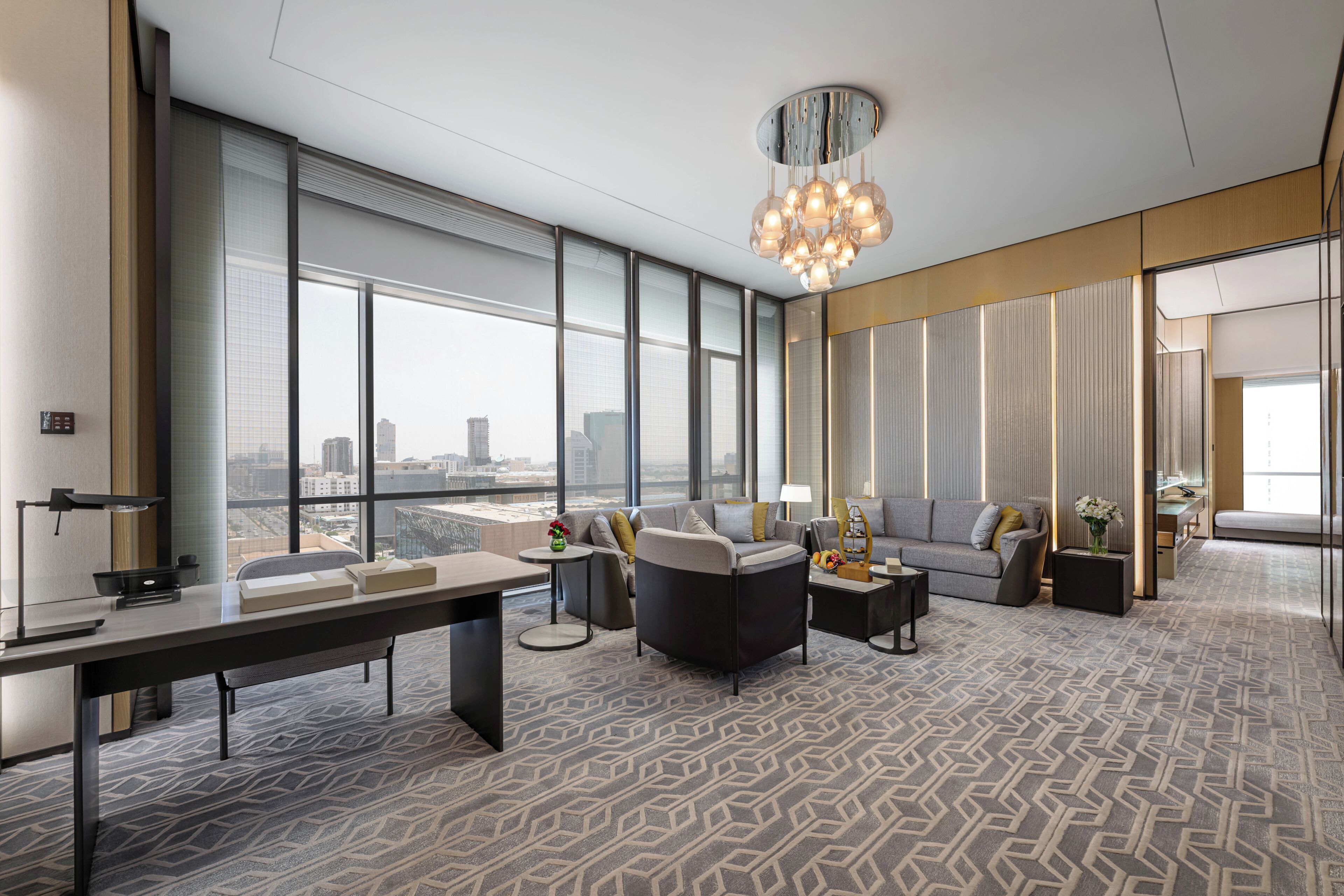 A spacious hotel room with a large desk, comfortable seating, a chandelier, and large windows offering a city view.