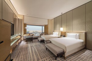 A hotel room with two beds, a desk, a chair, and a large window offering a city view.