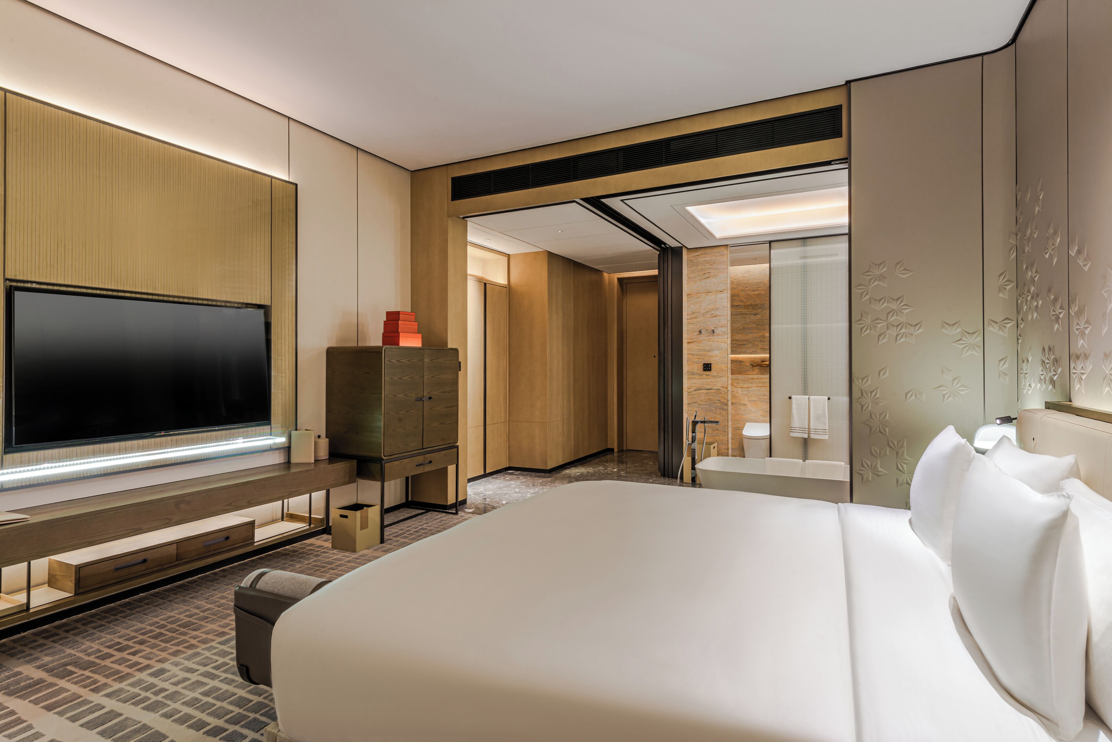 A modern hotel room with a large bed, a flat-screen TV, a wooden cabinet, and a bathroom with a mirror and towel rack.