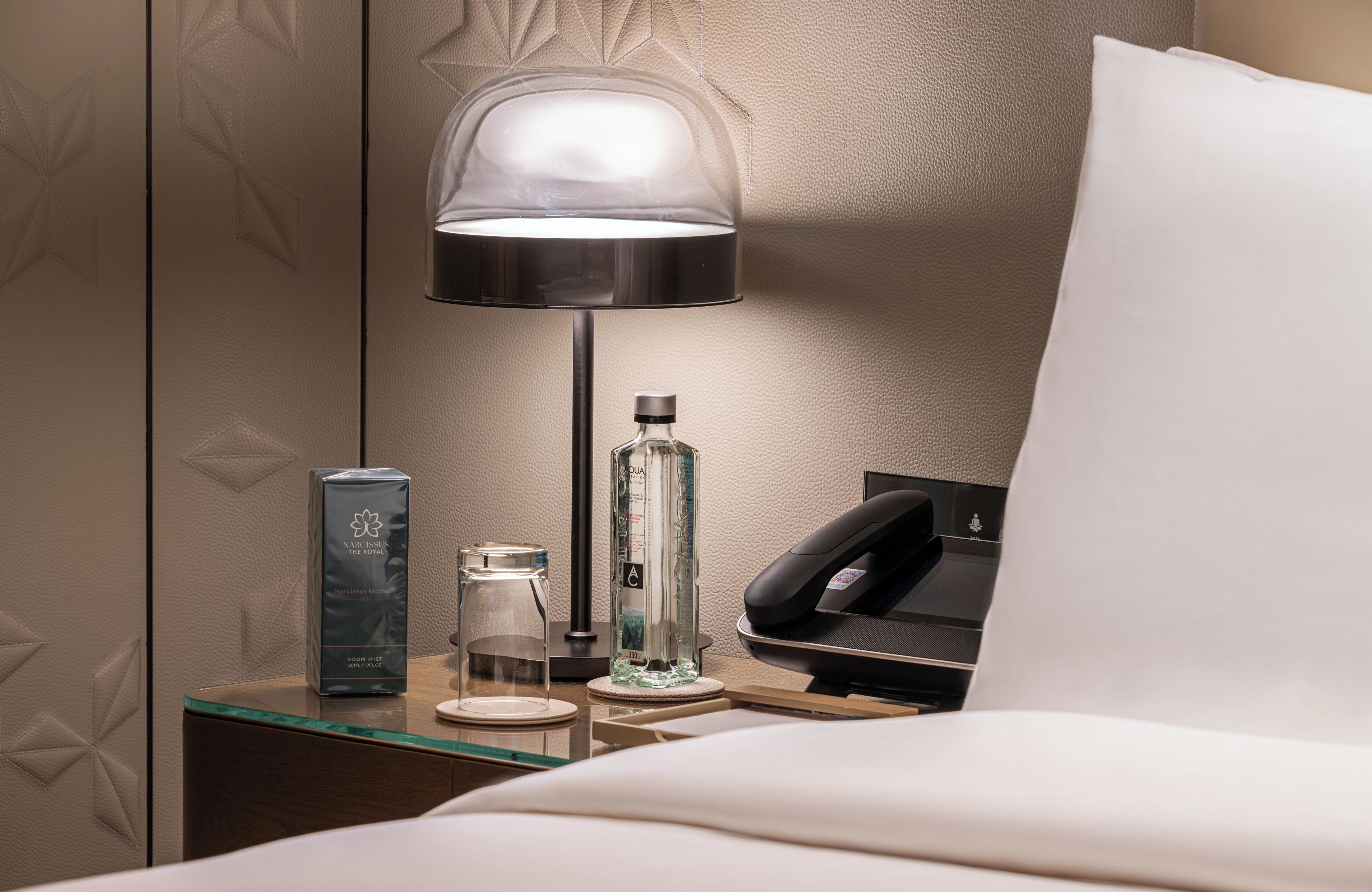 A hotel room with a neatly made bed, a bedside table with a lamp, a phone, a glass bottle, and a box of toiletries.