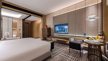 A modern hotel room with a large bed, a desk with a computer, a TV, and a small dining area with a table and chairs.