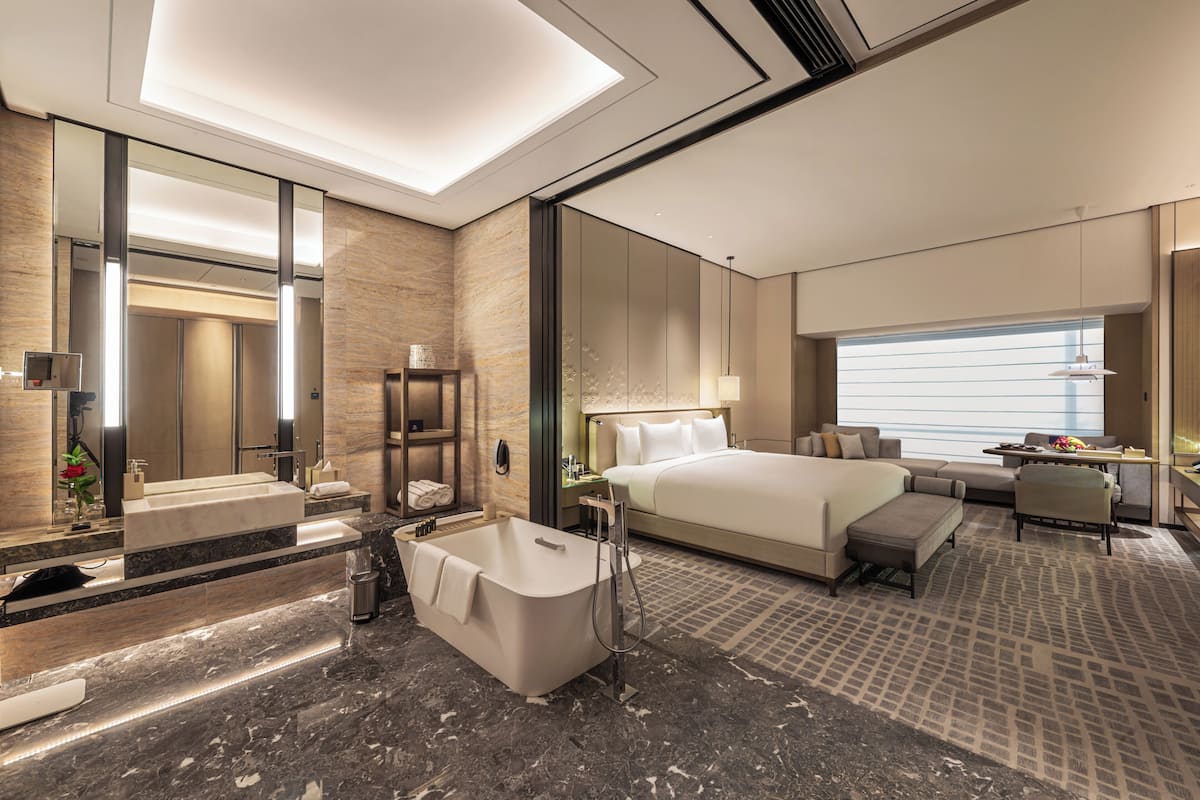 Une chambre d’hôtel moderne dotée d’un grand lit, d’un coin salon séparé, d’une salle de bain avec baignoire et d’une grande fenêtre offrant une vue dégagée.