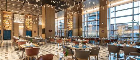 Un restaurante espacioso con techos altos, lámparas colgantes y ventanas amplias que ofrecen vistas de la ciudad.