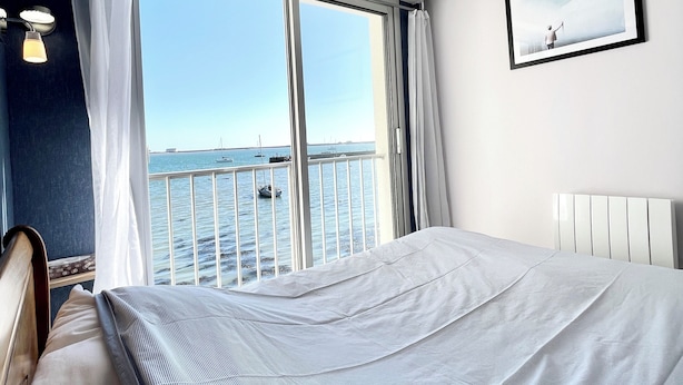 A hotel room with a large window offering a view of the sea and boats. There is a bed with a striped bedspread and a white headboard. - Pieds Dans L'eau T3 Top Cosy - Face à la mer (Port-Louis)