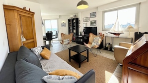 A living room with a sofa, armchairs, a coffee table, and a TV. There is a bookshelf, a dining area with a table and chairs, and a view of the sea through the window. - Pieds Dans L'eau T3 Top Cosy - Face à la mer (Port-Louis)