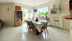 Un coin repas avec une table dressée pour quatre personnes, un espace cuisine avec un comptoir et des étagères, et une vue sur l’extérieur grâce à une grande fenêtre.