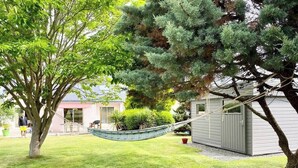 Un hamac est tendu entre deux arbres dans une cour verdoyante, avec un abri de jardin et une maison en arrière-plan.