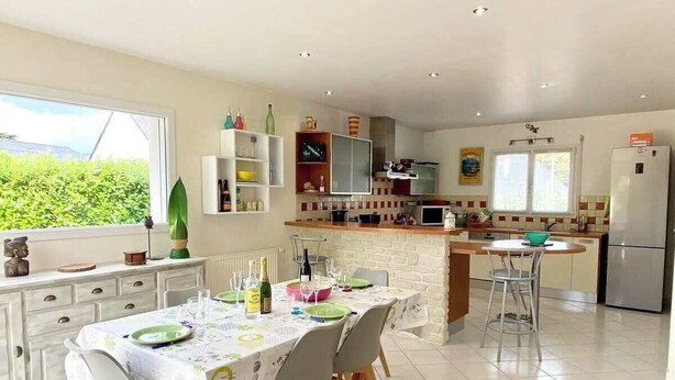 Une cuisine moderne avec des meubles blancs, un coin repas avec une table dressée pour quatre personnes, et une grande fenêtre offrant une vue sur la verdure environnante.