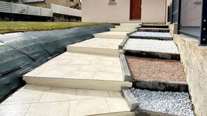 Un bâtiment à la façade beige, avec un toit foncé et une porte rouge. Des marches mènent à l’entrée, et une clôture avec un portail se trouve sur la droite.