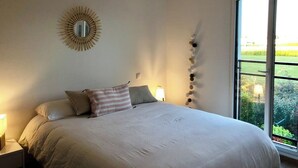 A bedroom with a large bed, a round mirror on the wall, a window with a view of greenery, and a ceiling light fixture.