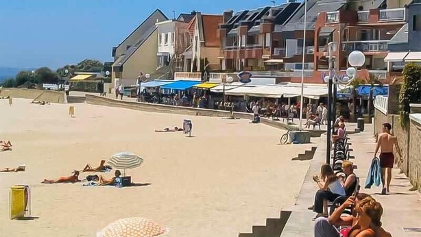 A beach with people sunbathing and walking, buildings with balconies, and a clear sky.