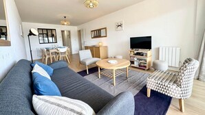 A modern living room with a grey sofa, a round coffee table, and a patterned armchair. There is a dining area with a table and chairs, and a television in the corner. - T3 Standing Vue Mer 180° Face Plage (Larmor-Plage)
