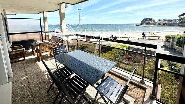 A balcony with a table and chairs, overlooking a beach and a coastal town. - T3 Standing Vue Mer 180° Face Plage (Larmor-Plage)