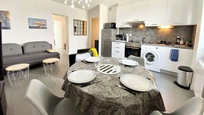 Eine moderne Küche mit weißen Schränken, einem eingebauten Backofen und einer Waschmaschine. Ein Essbereich mit Tisch für vier Personen und ein graues Sofa im Wohnzimmer.