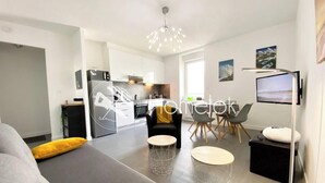 Ein modernes Wohnzimmer mit einer Couch, einem Essbereich mit Stühlen, einer Küche im Hintergrund und einem an der Wand befestigten Flachbildfernseher.