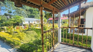 A balcony with a bamboo railing overlooking a lush garden and a building with a thatched roof. - Falcon Jungle Resort (Sarupeta)