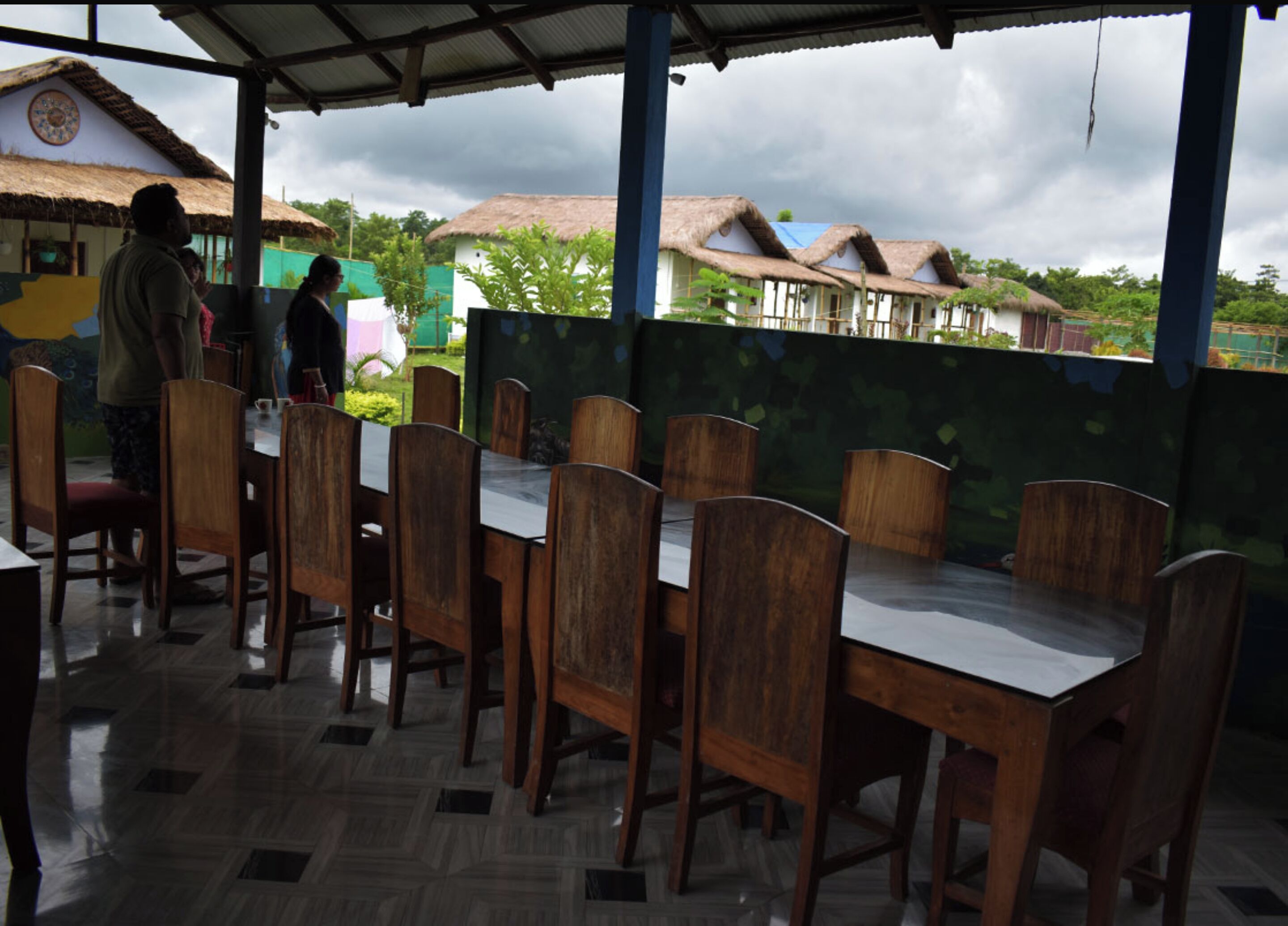 An outdoor dining area with wooden tables and chairs, a tiled floor, and a view of houses in the background.
