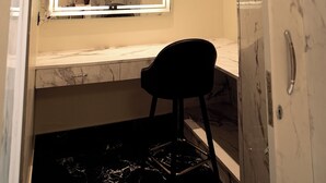 Un baño con tocador de mármol, espejo con marco iluminado, una silla negra y zona de ducha.