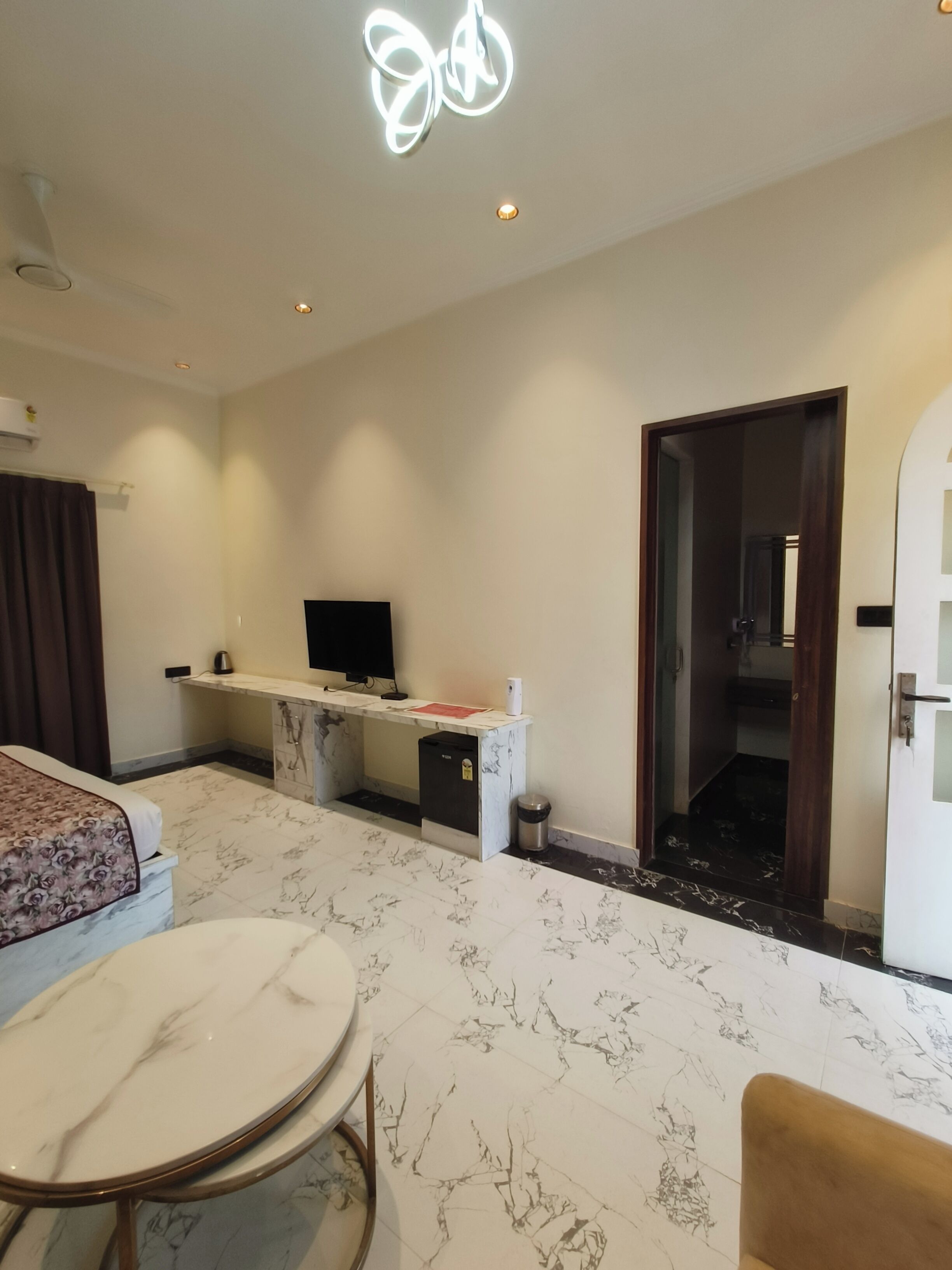 A hotel room with a bed, a desk with a TV, a small table, and a bathroom visible through an open door.