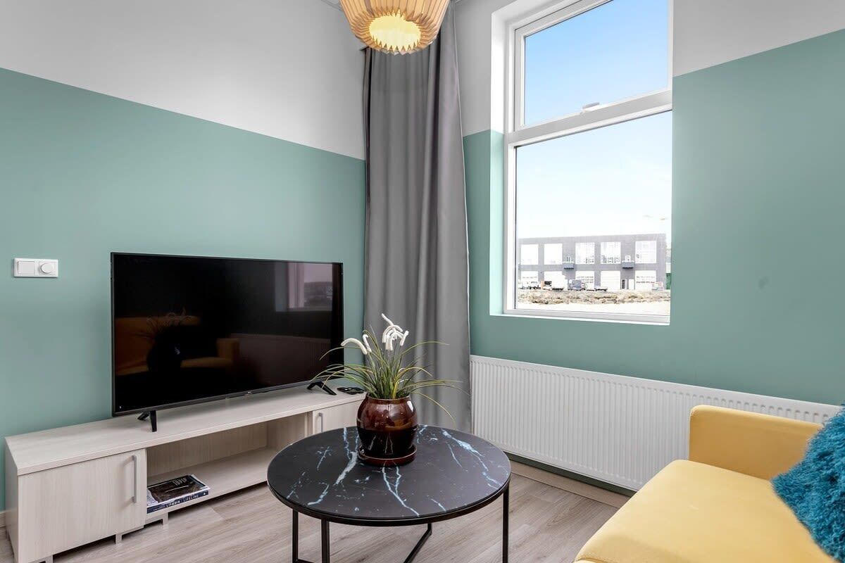 A modern living room with a flat-screen TV, a round coffee table, a yellow sofa, and a large window offering a view of a building outside.