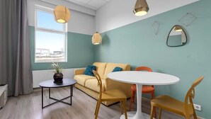 Ein modernes Wohnzimmer mit einer Couch, einem runden Tisch und Sesseln. Ein Fenster mit Vorhängen, eine Pflanze auf dem Tisch und ein Ausblick nach draußen.