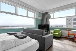Quarto de hotel moderno com uma cama grande, um sofá cinzento, uma mesa pequena com uma cadeira e grandes janelas com vista para a cidade.