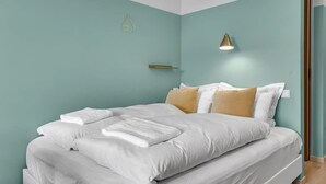 A neatly made bed with white linens and a single tan pillow, a bedside lamp, and a wall-mounted light fixture. - Icelandic Apartments- Economy Studio (Kópavogur)