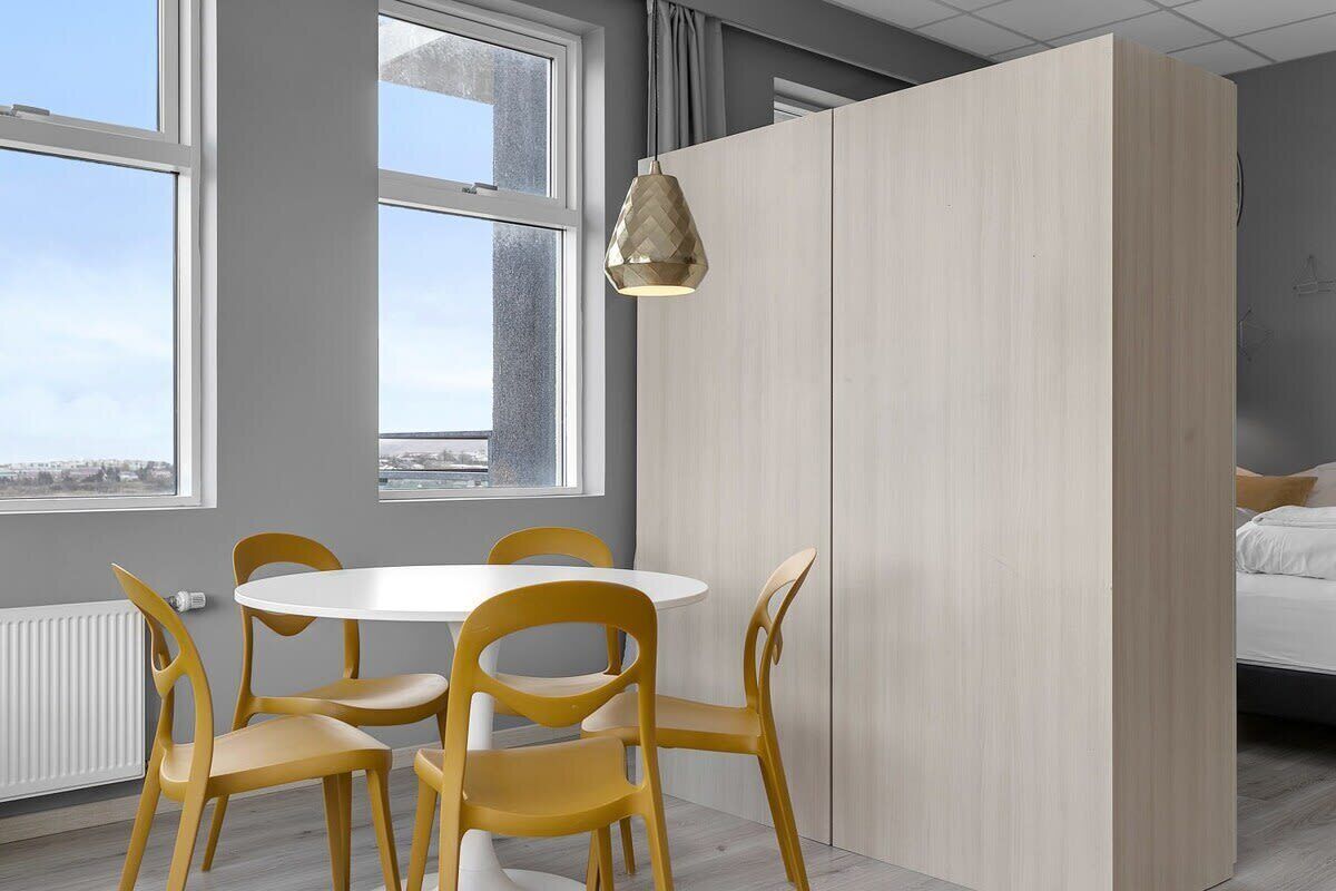 A modern room with a round table and yellow chairs, large windows, and a bed with white bedding.