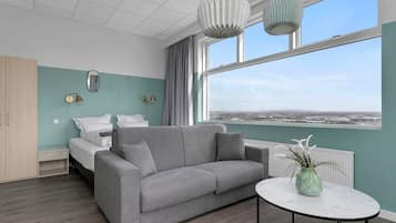 Une chambre moderne avec un canapé gris, un lit avec une literie blanche, une petite table avec une plante, et une vue sur la ville à travers de grandes fenêtres.