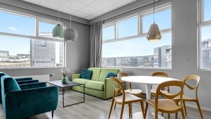 Un salon moderne avec un canapé vert, des fauteuils turquoise, une table à manger blanche et des chaises en bois. De grandes fenêtres offrent une vue sur la ville.