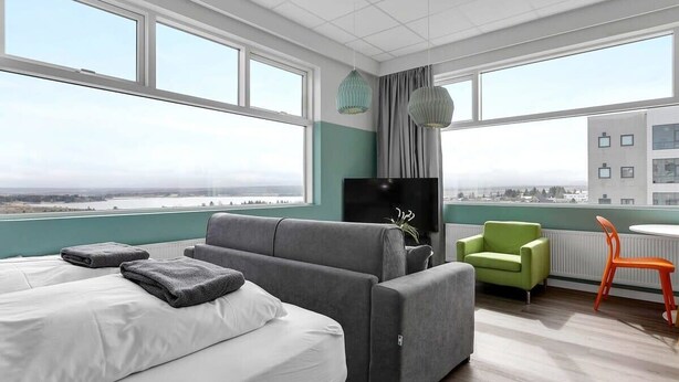 Una habitación de hotel moderna con una cama grande, un sofá gris, una mesita con una silla y ventanas amplias que dan a vistas de la ciudad.