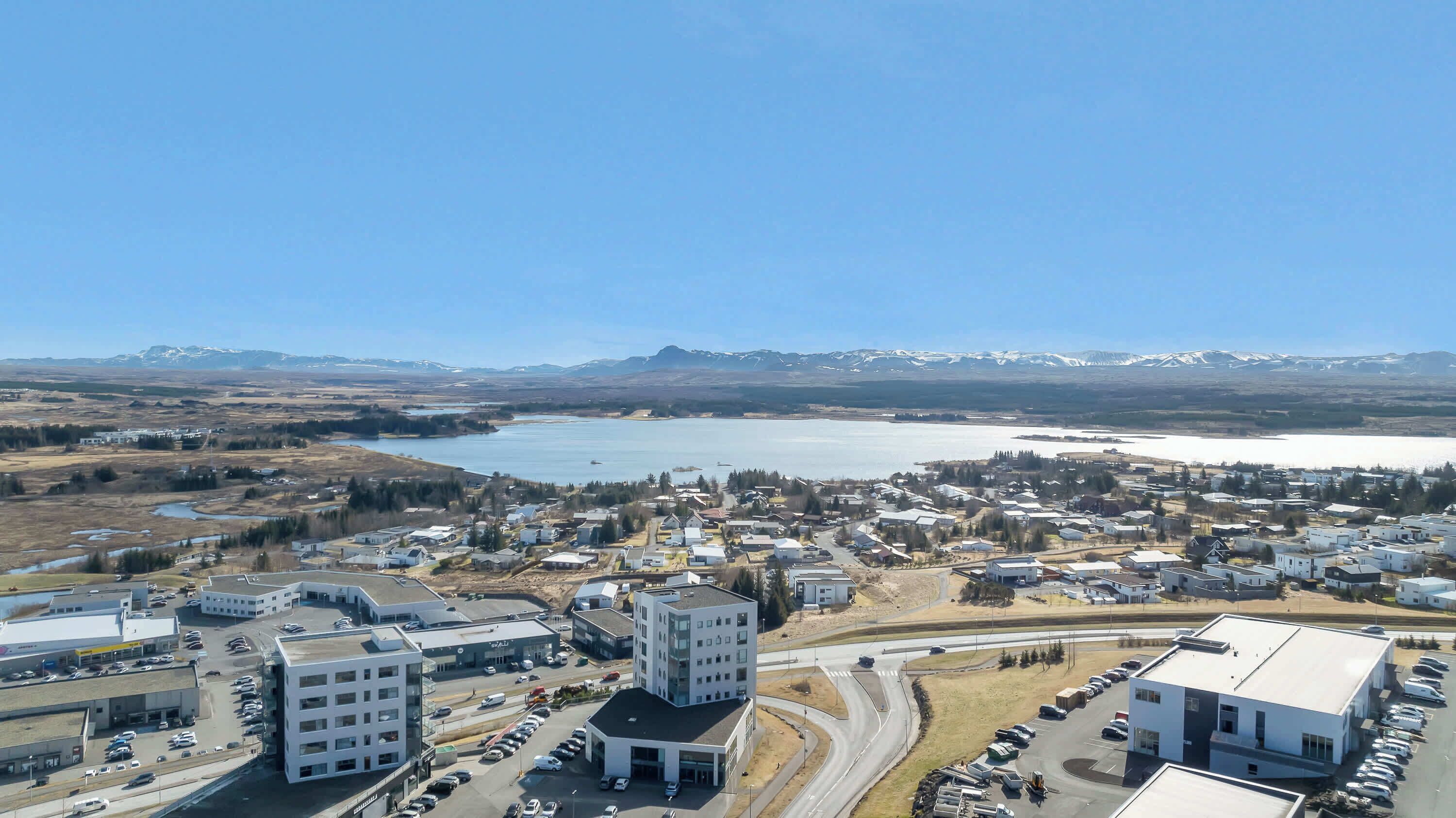 Une ville traversée par une rue principale, bordée de bâtiments, et donnant sur une vaste étendue d’eau, avec des montagnes en arrière-plan.