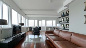 Un salón amplio con un sofá de cuero, una mesa de centro de vidrio y un televisor de pantalla plana sobre un mueble. Hay ventanas grandes que proporcionan luz natural y vistas al exterior.
