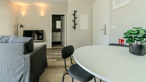 Uma sala de estar moderna com um sofá cinzento, uma mesa de jantar redonda e uma cadeira preta. Há uma pequena planta sobre a mesa, um cabideiro e uma porta que dá para uma cozinha.