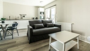 A modern living room with a sofa, a dining table with chairs, a kitchen area, and a radiator.