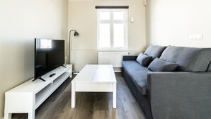 Una sala de estar con televisor, una mesa de centro blanca, un sofá gris y una ventana con persianas.