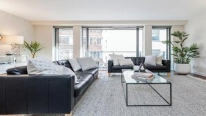 A modern living room with a black leather sofa, a glass coffee table, and a large window offering a view of buildings.