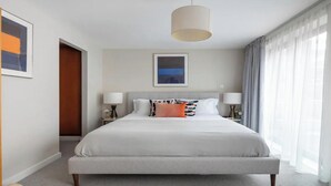 A modern hotel room with a large bed, two bedside tables with lamps, a framed artwork above the bed, and a view of the outdoors through sheer curtains.
