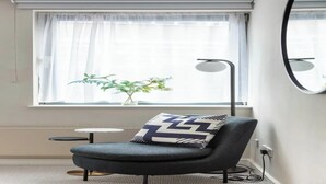 A modern room with a grey armchair, a small round table, a floor lamp, and a window with sheer curtains.