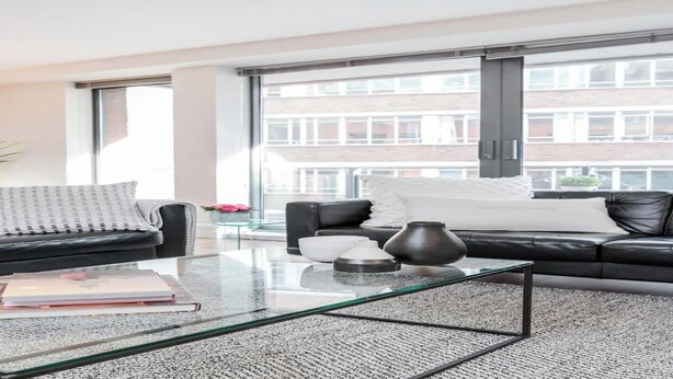 A modern living room with a glass coffee table, black leather sofa, and a large window offering a view of a building.