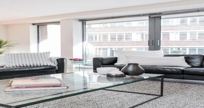 A modern living room with a glass coffee table, black leather sofa, and a large window offering a view of a building. - Modern 2-Bedroom, 2-Bathroom Condo in the Heart of London (London)