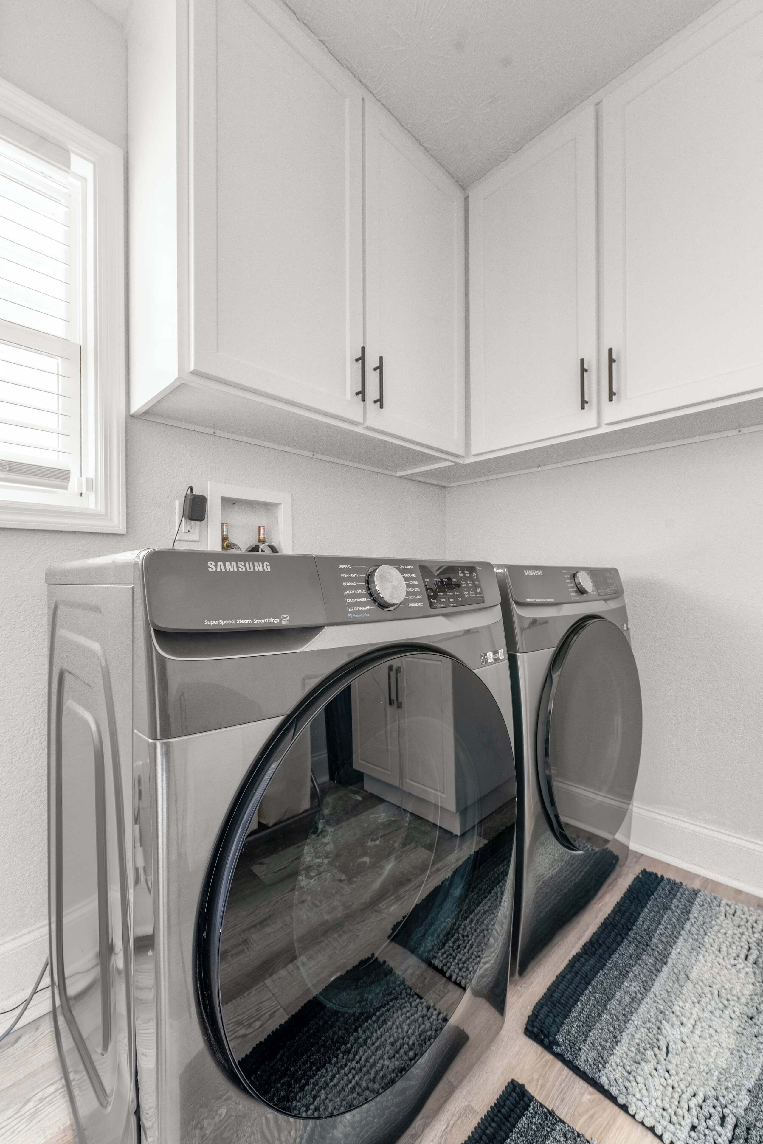 Une buanderie équipée de deux machines à laver Samsung SuperSpeed SteamWash, de meubles blancs et d’une fenêtre.