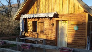 Puinen mökki, jossa on kalteva katto, etuovi ja pieni kuisti penkkeineen.
