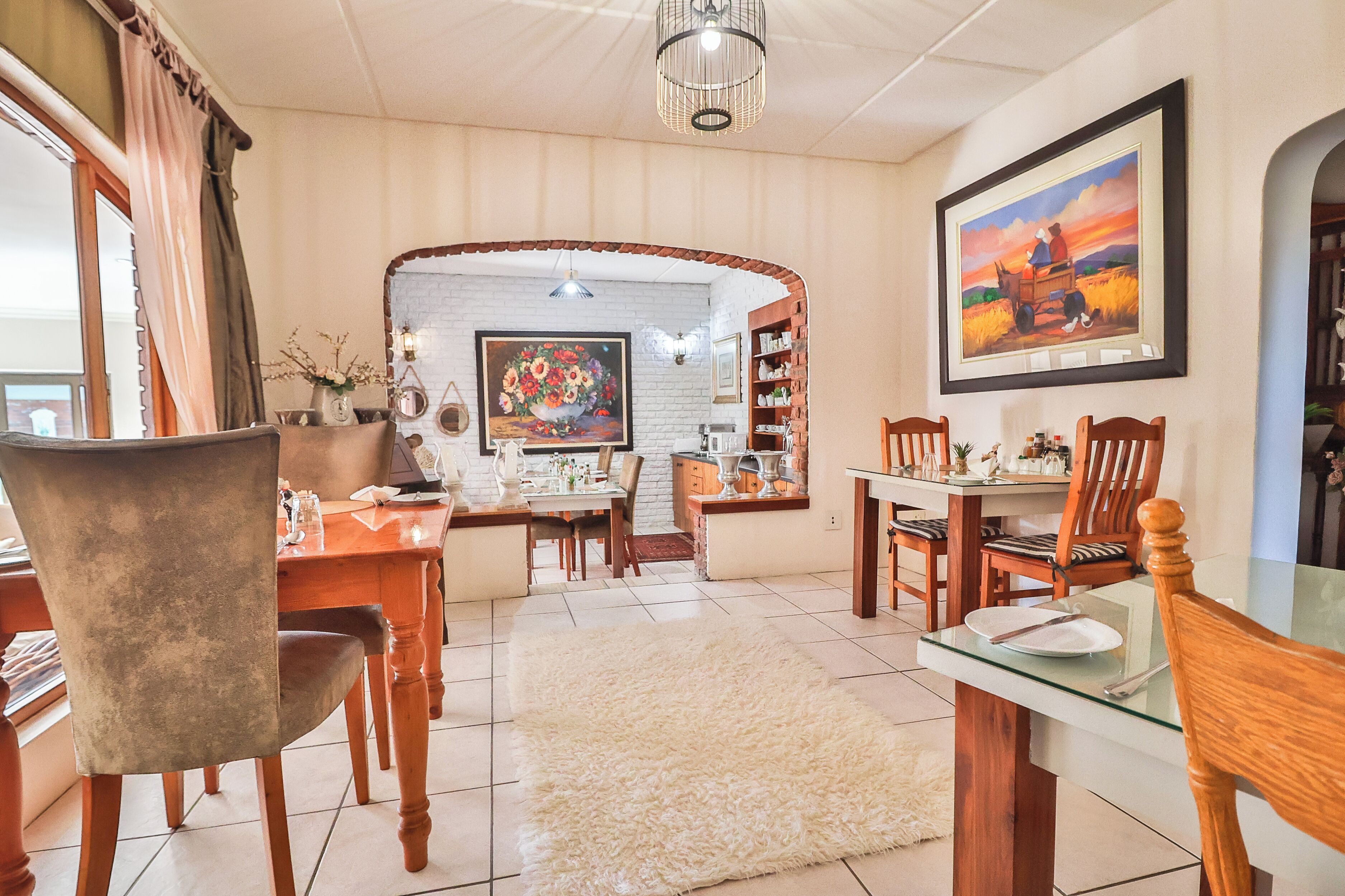 A dining area with wooden chairs, a glass table, and a white rug. There is a painting on the wall and a bookshelf with various items.