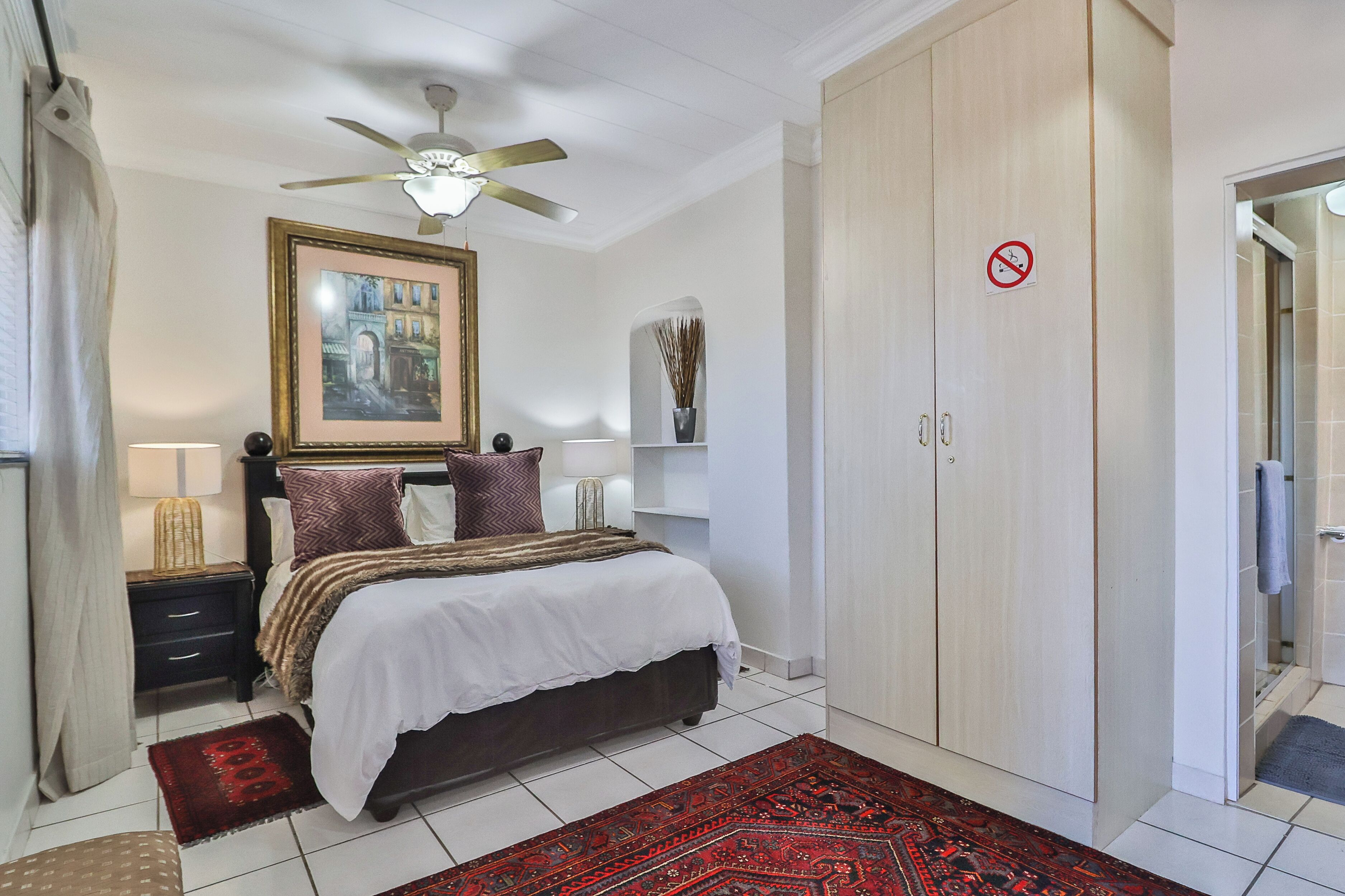 A bedroom with a bed, nightstands, a ceiling fan, a wardrobe, and a bathroom door.