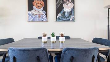 Un comedor con una mesa de madera, sillas tapizadas en azul y dos cuadros enmarcados que representan un perro y un gato con vestimenta histórica.