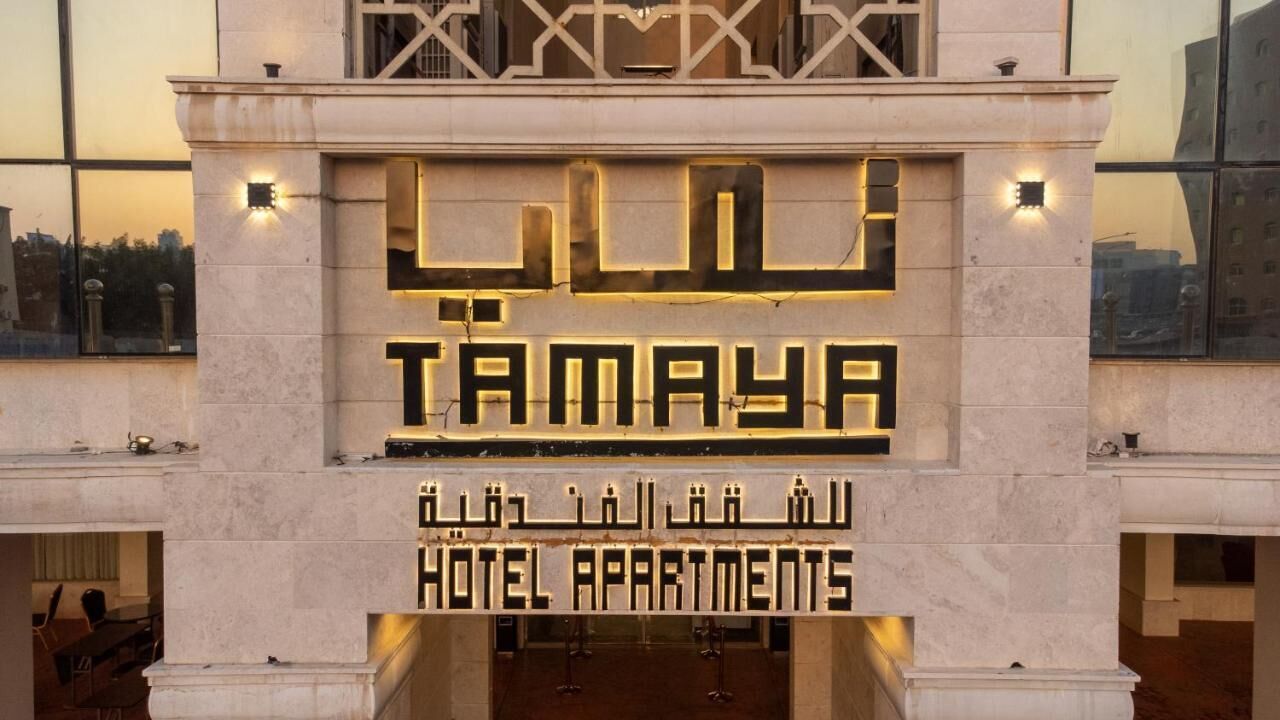 Entrada de un hotel con el nombre "Tamaya" y "Hotel Apartments" en árabe e inglés.