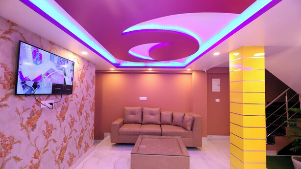 A modern living room with a sofa and ottoman, a flat-screen TV mounted on the wall, and a staircase with a yellow accent wall. - Jupiter Plaza (New Delhi)